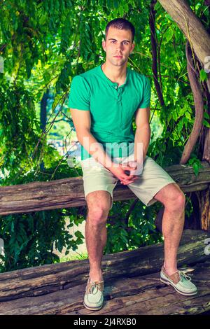 Un jeune homme se détendant dehors. Portant un t-shirt à manches courtes vert Henley, un short jaune clair, des chaussures décontractées en cuir, un jeune homme beau est assis sur le t Banque D'Images