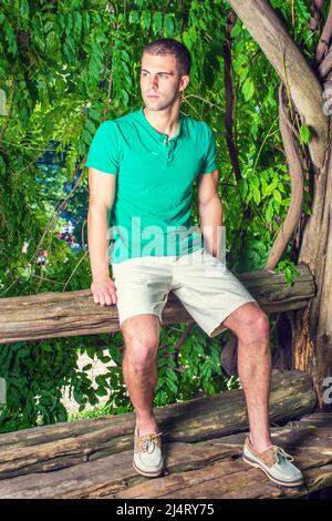 Un jeune homme se détendant dehors. Portant un t-shirt à manches courtes vert Henley, un short jaune clair, des chaussures décontractées en cuir, un jeune homme beau est assis sur le t Banque D'Images