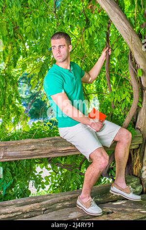 Vous vous demandez. Portant une chemise à manches courtes, un short, des chaussures décontractées en cuir, tenant un livre rouge, un jeune homme assis sur un tronc d'arbre, regardant autour. Concept de i Banque D'Images