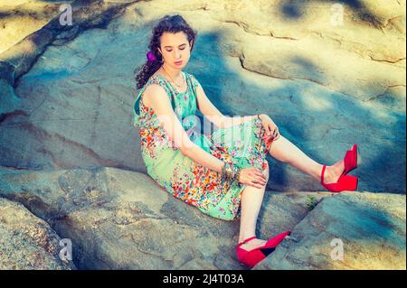 Mode d'été. Habillez-vous dans une robe longue sans manches, des chaussures de sandales rouges, un bracelet à grosse chaîne, une belle adolescente est assise sur un grand rochers Banque D'Images