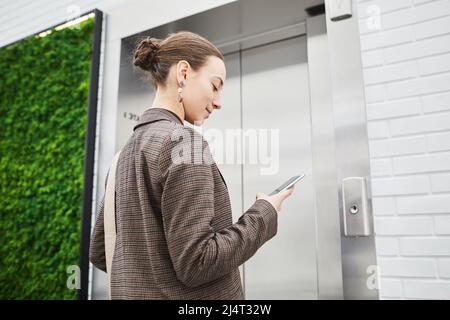 Photo sous angle minimal d'une jeune femme d'affaires qui attend un ascenseur dans un immeuble de bureaux moderne et qui utilise le téléphone et l'espace de copie Banque D'Images