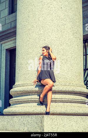 Étudiant. Habille en noir, haut sans manches, pantalon court, talons hauts, une jeune femme avec de longs cheveux bouclés est debout contre une colonne dehors Banque D'Images