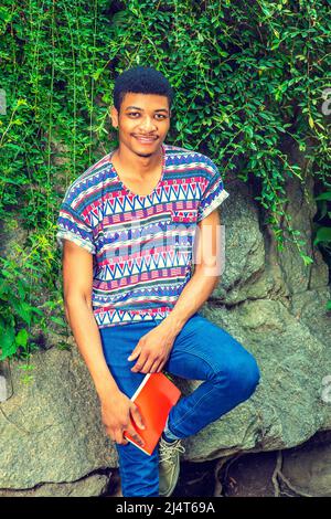 Portrait de Black College étudiant. Portant une chemise à manches courtes, sans col, à motif coloré, un Jean bleu, tenant un livre rouge, un jeune homme est assis Banque D'Images