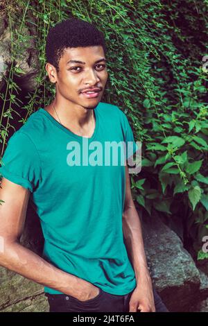 Portrait de jeune homme noir. Portant un t-shirt vert à manches courtes et col en V, un pantalon noir, une main dans la poche, un jeune homme charmant se penche contre roc Banque D'Images