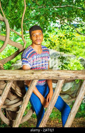 Jeune Noir vous attend. Portant une chemise à motif coloré, un Jean bleu, des baskets, un collier, un jeune homme beau est assis derrière un fen bois Banque D'Images