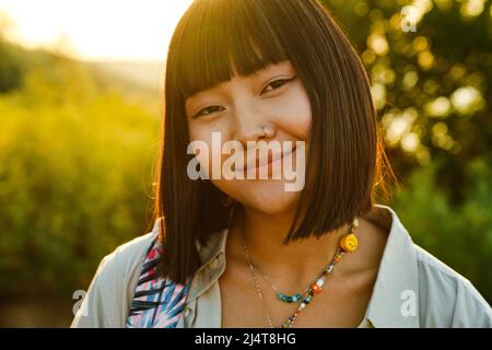 Jeune femme asiatique avec piercing souriant et souriant à la caméra à l'extérieur Banque D'Images