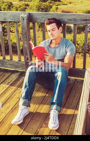 Homme lisant à l'extérieur. Portant un T-shirt gris, un Jean, des baskets blanches, un jeune homme beau est assis sur le sol en bois, dos contre la clôture dans un rempart Banque D'Images