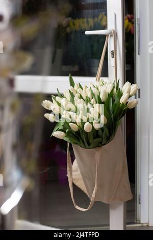 Composition avec de belles fleurs fraîches tulipes blanches dans un sac ECO suspendu sur la poignée de porte. Modèle à imprimer. Shopping de printemps. Banque D'Images