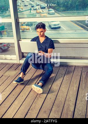 Étudiant occupé. Portant un t-shirt à col en V noir, un Jean bleu, des baskets, une montre-bracelet, un jeune homme assis sur un sol en bois, contre un mur de verre, travaillant Banque D'Images