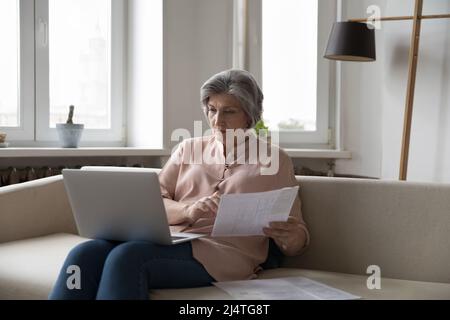 Une femme âgée sérieuse est assise sur un canapé avec un ordinateur portable, analyse des papiers Banque D'Images