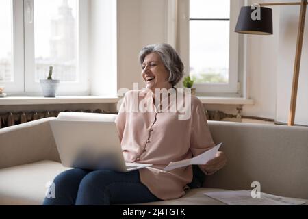 Joyeux rire grand-mère s'asseoir sur un canapé avec un ordinateur portable et des factures Banque D'Images