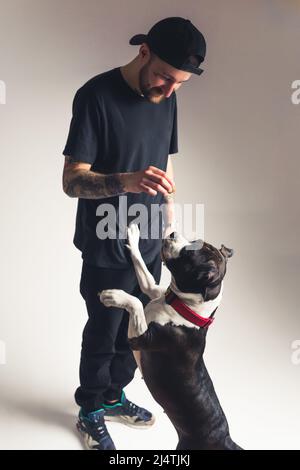 Relation entre le concept animal et humain. Smiley caucasien gars avec tatouages jouant avec son chien d'origine dans un studio sur fond gris. Tir vertical. Photo de haute qualité Banque D'Images