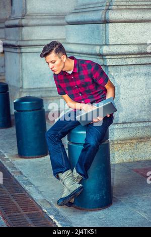 Distraction. Portant une chemise à manches courtes et à motifs, un Jean, des chaussures de démarrage, tenant un ordinateur portable, un jeune homme est assis sur un pilier en métal, regardant Banque D'Images