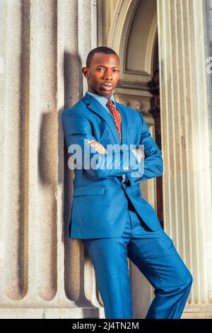 Gestion des affaires Robe officiellement en costume bleu, sous-chemise à motifs, cravate, coupe courte, bras croisés, un jeune homme noir est debout près d'une colonne outsi Banque D'Images