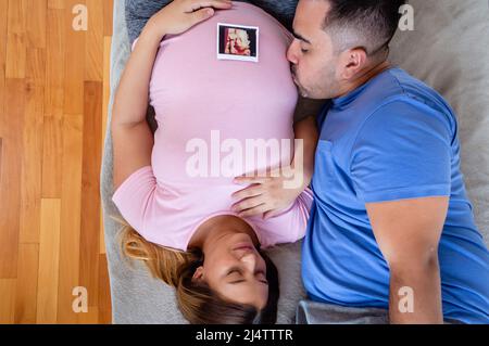 le jeune homme caucasien embrasse sa petite amie enceinte sur le ventre, couple hétérosexuel enceinte allongé sur le lit, concept de mode de vie. Banque D'Images