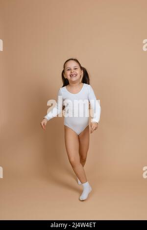 Portrait de jeune belle heureuse fille souriante avec de longs cheveux foncés en maillot de bain blanc pour la gymnastique et des chaussettes debout sur fond marron, posant Banque D'Images