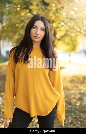 une jeune femme sérieuse dans un chandail d'orange se repose dans un parc avec des arbres jaunes en arrière-plan. belle dame bénéficie d'un temps chaud et ensoleillé à autum Banque D'Images