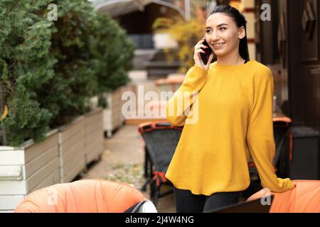 Femme d'affaires parlant sur un téléphone cellulaire, debout à la table d'un café de rue, une fille brune sourit et parle à un client sur un mobile, la technologie A. Banque D'Images