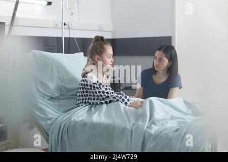 Mal blessée, petite fille portant un col de l'utérus parlant à une mère inquiète et inquiète pendant qu'elle est assise sur le lit d'hôpital. Blessé petite fille portant un collier médical discutant avec une femme attentionnée. Banque D'Images