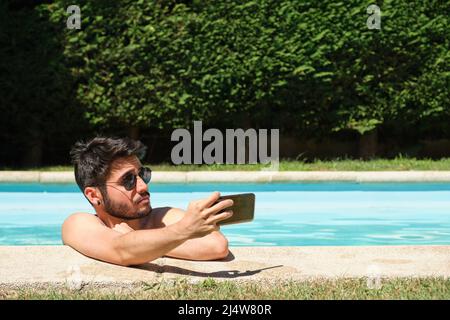 Jeune homme portant des lunettes de soleil, prenant un selfie dans une piscine. Banque D'Images