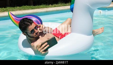 Jeune homme portant des lunettes de soleil prenant un selfie sur un anneau gonflable unicorn. Banque D'Images
