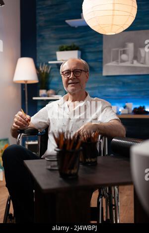 Portrait d'un homme souriant aux cheveux gris plus vieux posant un crayon confiant devant la table avec des outils de dessin professionnels en studio d'art. Professeur d'art à la retraite en fauteuil roulant se sentant créatif. Banque D'Images