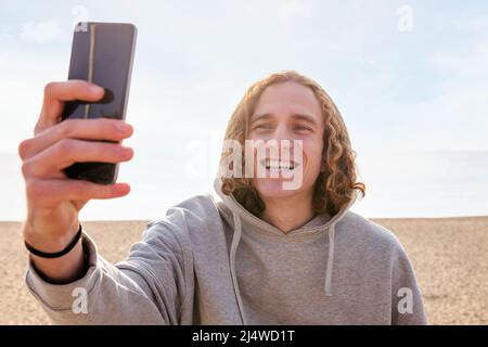 souriant homme caucasien dans un sweat à capuche prenant un selfie avec son téléphone à côté de la plage, concept de style de vie et de technologie Banque D'Images