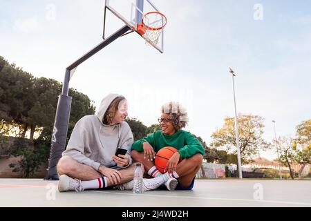 couple d'amis riant et s'amusant assis sur le terrain après un entraînement de basket-ball dans un parc de ville, concept d'amitié et de sport urbain dans le th Banque D'Images