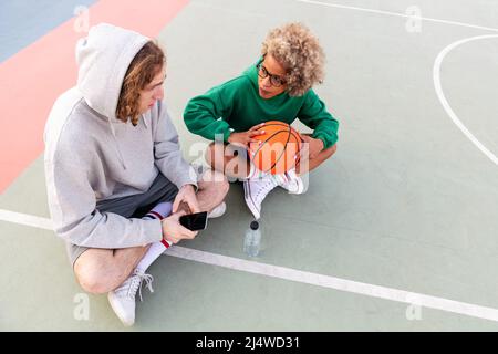 couple d'amis discutant et partageant un bon moment assis sur le terrain après un match de basket-ball dans un parc de ville, concept d'amitié et de sport urbain Banque D'Images