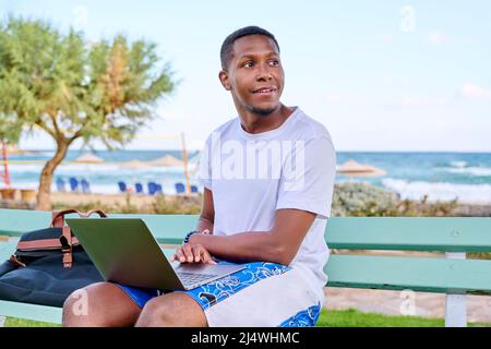 Jeune homme souriant indépendant se détendant sur la plage, assis sur le banc à l'aide d'un ordinateur portable Banque D'Images