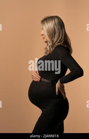 Femme enceinte en robe noire serrée posant en studio sur un fond beige Banque D'Images