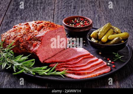 pastrami rôti de bœuf tranché sur une assiette noire avec romarin frais, thymes et cornichons sur une table sombre, vue horizontale depuis le dessus Banque D'Images