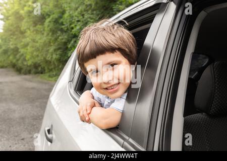 Un petit garçon souriant se déplace en voiture, regarde par la fenêtre ouverte, s'amuser, profiter de la conduite en voiture, à l'extérieur en été Banque D'Images