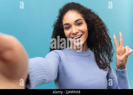Jeune femme afro-américaine joyeuse en chandail lavande prenant selfie, belle étudiante multiraciale tenant la main dans le geste de victoire, souriant et regarde l'appareil photo isolé sur bleu pastel Banque D'Images