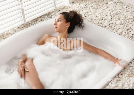 Femme millénaire rêveuse se détendant dans un bain chaud à bulles, regardant la fenêtre et méditant à la maison, espace libre Banque D'Images