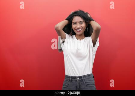 Portrait d'une jeune fille indienne confuse couvrant et tirant les cheveux avec les paumes et faisant le visage étrange. Isolé sur fond rouge. Banque D'Images