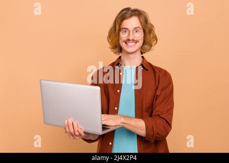 Portrait d'un beau gars à cheveux rouges, enjoué et enjoué, avec un ordinateur portable isolé sur un fond beige pastel Banque D'Images