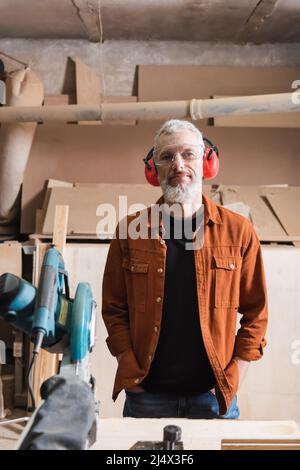 concepteur de meubles matures dans des lunettes et des casques antibruit debout avec les mains dans les poches dans l'atelier Banque D'Images