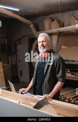 concepteur de meubles barbus regardant la caméra près d'un ordinateur portable dans un studio de travail du bois Banque D'Images