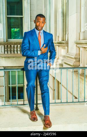 Robe de costume bleu, cravate, chaussures en cuir, mettre la main sur la poitrine, un jeune homme d'affaires noir assis sur la main courante dans un immeuble de bureau de style vintage, con Banque D'Images