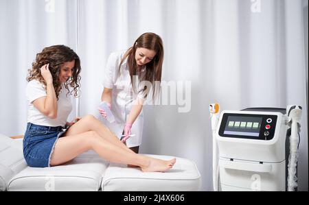 Une femme heureuse assise sur un lit de jour près de l'appareil d'épilation à diode laser tout en ayant une procédure d'épilation en clinique. Un cosmétologue souriant en gants stériles applique du gel sur la jambe avant l'épilation. Banque D'Images