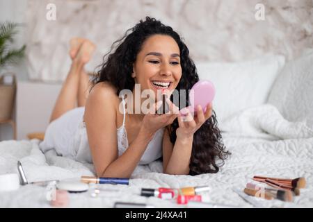 Gaie jeune caucasienne brune femme avec de longs cheveux bouclés en pyjama se trouve sur le lit et met le rouge à lèvres Banque D'Images