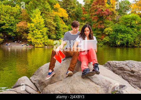 Jeune couple étudiant à l'extérieur près d'un lac, gars portant un t-shirt rayé, pantalon marron, baskets, tenant un livre, robe de fille en haut blanc, pantalon rouge, Banque D'Images