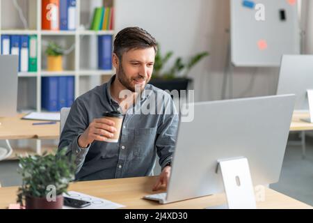 Heureux homme d'affaires mature travaillant sur ordinateur et de boire du café à emporter, dactylographiant sur le clavier, travaillant en ligne dans le bureau Banque D'Images
