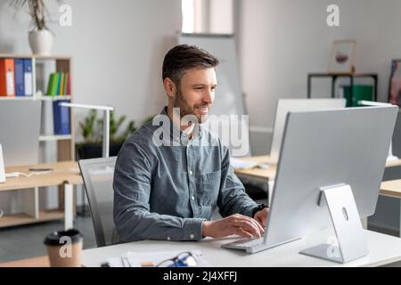 Homme d'affaires joyeux d'âge moyen assis à la table de travail, dactylographiant sur le clavier d'ordinateur, travaillant sur la recherche de marketing Banque D'Images
