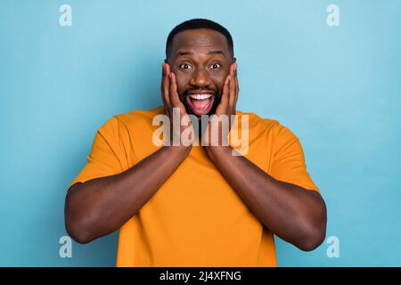 Photo de funky choqué gars habillé t-shirt orange souriant bras joues isolé couleur bleu fond Banque D'Images