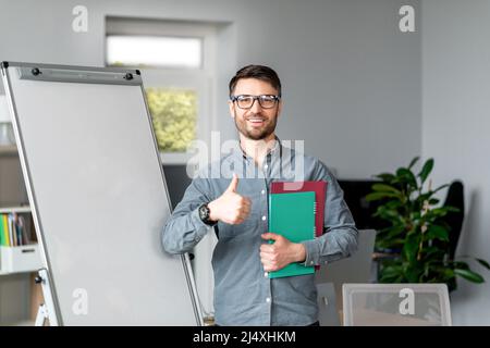 Un bon tuteur d'homme d'âge moyen qui donne un webinaire ou une conférence aux étudiants, en montrant le pouce vers le haut, en faisant une maquette sur un tableau noir vide Banque D'Images