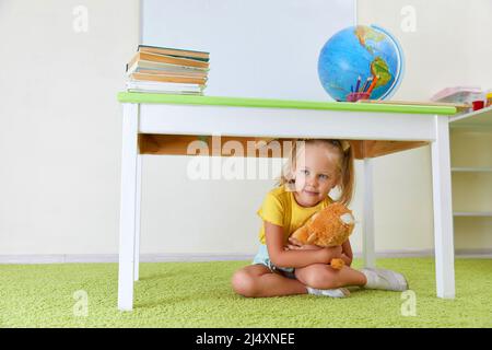 Fille effrayée se faisant cacher sous une table assise sur un sol Banque D'Images