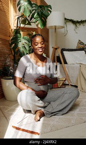 Femme heureuse africaine assise sur le sol dans la chambre à coucher et touchant son ventre enceinte, elle appréciant sa grossesse Banque D'Images