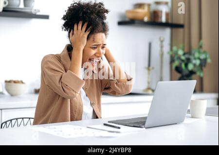 Frustré contrarié afro-américain brunette fille, indépendant, directeur, assis à la table dans la cuisine, utilise l'ordinateur portable tandis que le travail à distance, choqué par les nouvelles ou le message, a subi des pertes financières, cri Banque D'Images